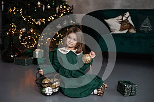 Smiling little girl in dress sit on floor with gift and Xmas balls toy by Christmas tree with glowing garland lights. Beautiful sm