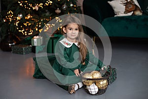 Smiling little girl in dress sit on floor with gift and Xmas balls toy by Christmas tree with glowing garland lights. Beautiful sm