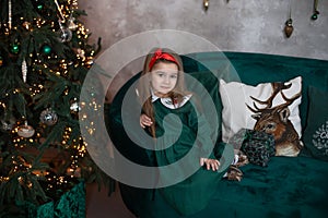 Smiling little girl in dress sit with gift on sofa by Christmas tree with glowing garland lights. Beautiful small girl in living r