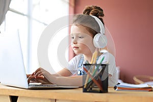 Smiling little girl in headphones handwrite study online using laptop at home