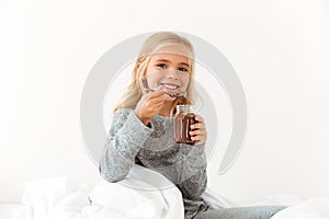 Smiling little girl holding bank of sweet chocolate hazelnut spread, looking at camera while sitting in bed