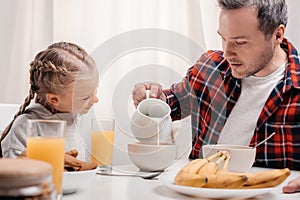 smiling little girl looking at father pouring milk into bowl with flakes