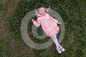 Smiling little girl lying on the grass. Preschool girl in pink dress lay down to rest on the green meadow. Top view
