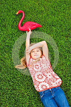 Smiling little girl lying on green grass with pink flamingo
