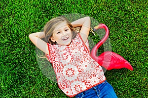 Smiling little girl lying on green grass with pink flamingo