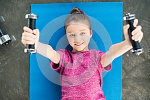 Smiling little girl lying on mat and exercising with dumbbells