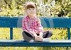 A smiling little girl is sitting on a bench on a sunny summer day in the park. A cute little girl is sitting in a lotus position