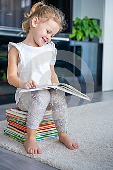 Smiling Little girl is sitting on stack of children& x27;s books and leafing through a book with fairy tales