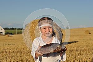 A smiling little girl in slavic traditional ornamented chemise holding a loaf of a rye bread in the harvested filed