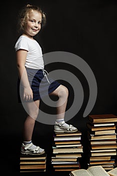 A smiling little girl steps in stacks of books. Knowledge and education. Back to school. Black background. Full height