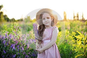 Smiling little girl in straw hat holds lupine flower in her hands at sunset. Child Girl in field of lupines. Childhood concept. Co