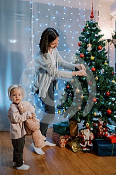 Smiling little girl with a teddy bear in an embrace stands near her mother hanging colorful balls on the Christmas tree