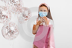 Smiling little girl wearing a medical protective mask on her face celebrating her birthday with balloons and shopping bags on a