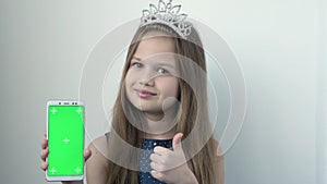 Smiling little girl wearing a princess crown and blue festive dress shows phone with green screen. Chroma key green