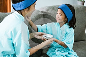smiling little girl wearing a towel applies a black clay face mask to her mother& x27;s face