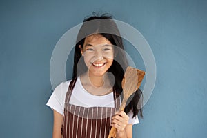 Smiling long brunette hair 10s Asian girl in brown stripped apron holding wood spatula for home cooking food with blue background