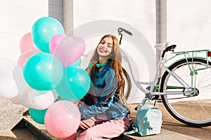 Smiling long-haired girl sitting on the steps with blue and pink helium balloons ready for bike trip around the town