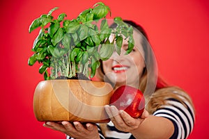 Smiling long-haired plump woman in striped shirt showing to camera green basil plant pot and fresh apple in red studio