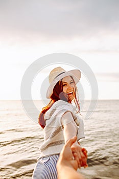 Smiling long haired woman in a hat holding boyfriends hand along empty ocean beach sand at sunset . Follow me concept