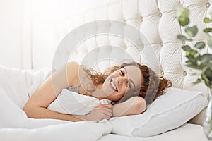 Smiling lovely young woman wakes up in morning, has gladful expression, lies on white bedclothes in bed, poses at camera with sati