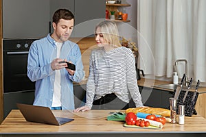 Smiling loving couple cooking salad together while standing on a kitchen at home and using mobile phone and laptop