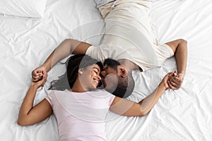 Smiling loving young african american lady and guy enjoy romantic moment lie on comfortable bed in bedroom, top view