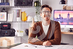 Smiling Male Architect In Office Working At Desk With Wooden Model Of Building