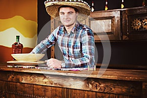 Smiling male bartender in a sombrero leaned on bar counter in Me