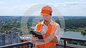 Smiling male builder in helmet working the roof of construction site while typing on digital tablet. Business, building