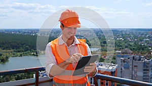 Smiling male builder in helmet working the roof of construction site while typing on digital tablet. Business, building
