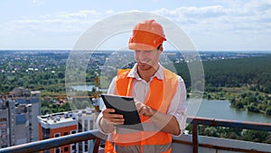 Smiling male builder in helmet working the roof of construction site while typing on digital tablet. Business, building