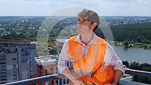 Smiling male builder looking on the construction site while holding helmet and drawing. Business, building, industry