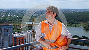 Smiling male builder looking on the construction site while holding helmet and drawing. Business, building, industry