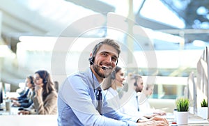 Smiling male call-center operator with headphones sitting at modern office with collegues on the backgroung