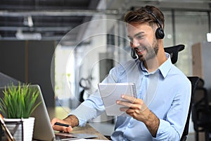 Smiling male call-center operator with headphones sitting at modern office, consulting online information in a laptop