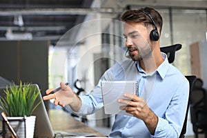 Smiling male call-center operator with headphones sitting at modern office, consulting online information in a laptop