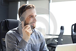 Smiling male call-center operator with headphones sitting at modern office, consulting online information in a laptop