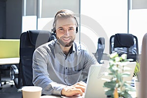 Smiling male call-center operator with headphones sitting at modern office, consulting online information in a laptop