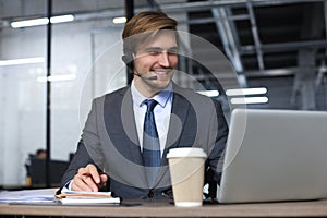 Smiling male call-center operator with headphones sitting at modern office, consulting online information in a laptop