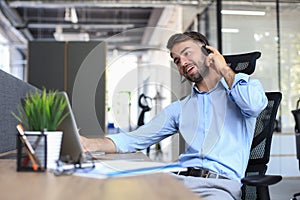 Smiling male call-center operator with headphones sitting at modern office, consulting online information in a laptop