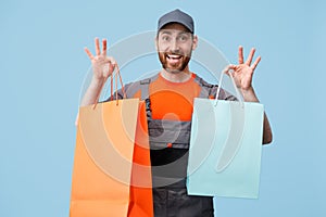 Smiling male courier in uniform holding shopping bags on blue studio background