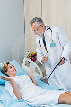 smiling male doctor with stethoscope over neck holding clipboard and talking to female patient in hospital