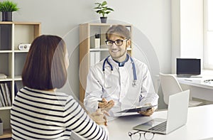 Smiling male doctor talking to female patient sitting at desk at clinic or hospital