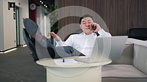 Smiling male employee resting at workplace and talking on modern smartphone