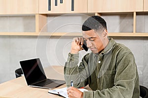 Smiling male employee using laptop for remote work, sits at desk in home office, talking on smartphone and taking notes