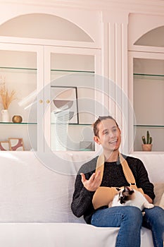 Smiling male with fractured hand in sling sitting with cat in living room at apartment during daytime