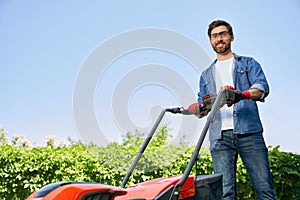 Smiling male gardener in protective glasses using lawn mower, while gardening in sunny day.
