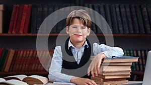 Smiling male kid pupil posing at public library desk with stack of paper books enjoy learning