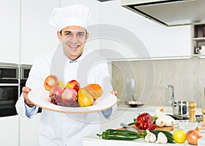 Smiling male kitchener in uniform is posing with plate of fruits in the kitchen
