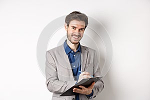 Smiling male manager in suit writing on clipboard, taking notes at office meeting, standing on white background